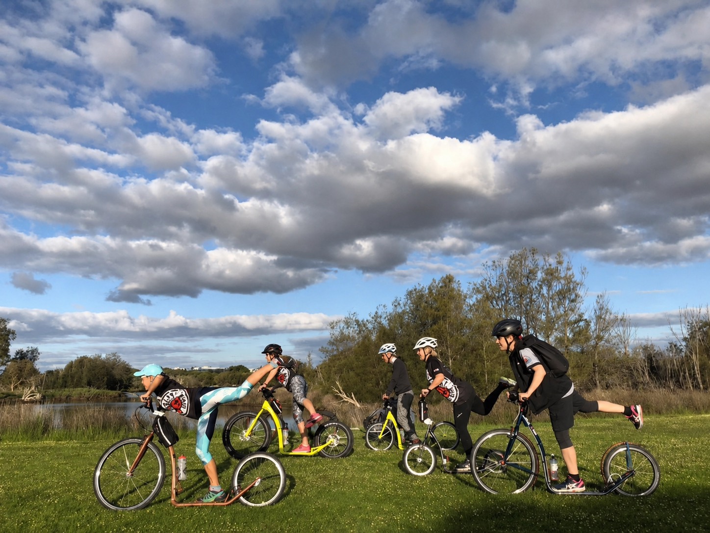 Five riders on footbikes on grass beside a lake near Perth